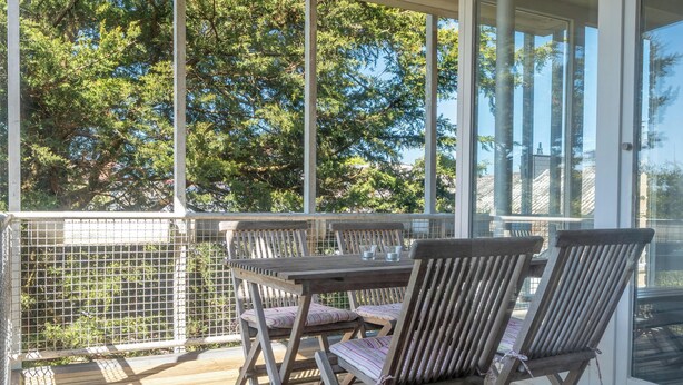 A covered outdoor seating area with a table and chairs, surrounded by a metal fence, overlooking a wooded area. - Vacation apartment Hafenperle (Laboe)