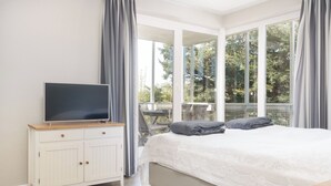 A bedroom with a bed, a TV on a dresser, and a balcony with a view of trees. - Vacation apartment Hafenperle (Laboe)