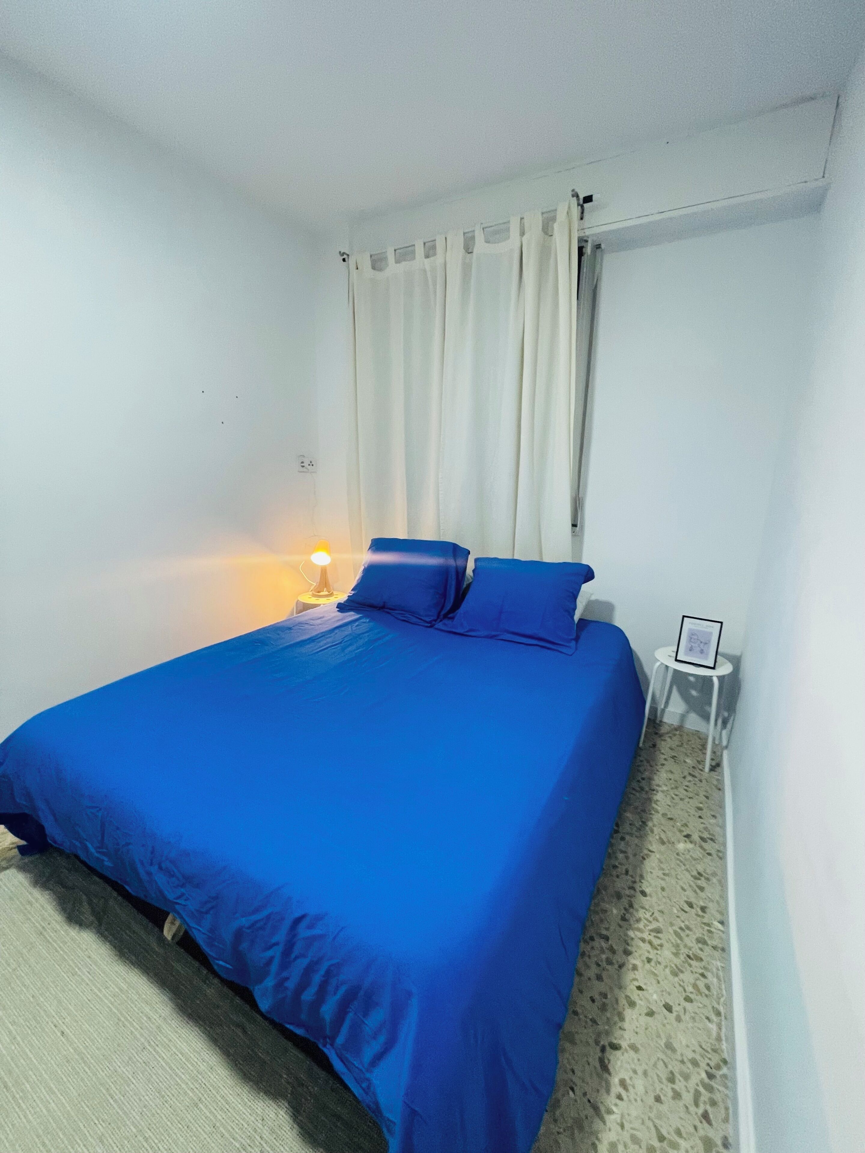 A bedroom with a bed, a nightstand, a lamp, and a small table with a picture frame.