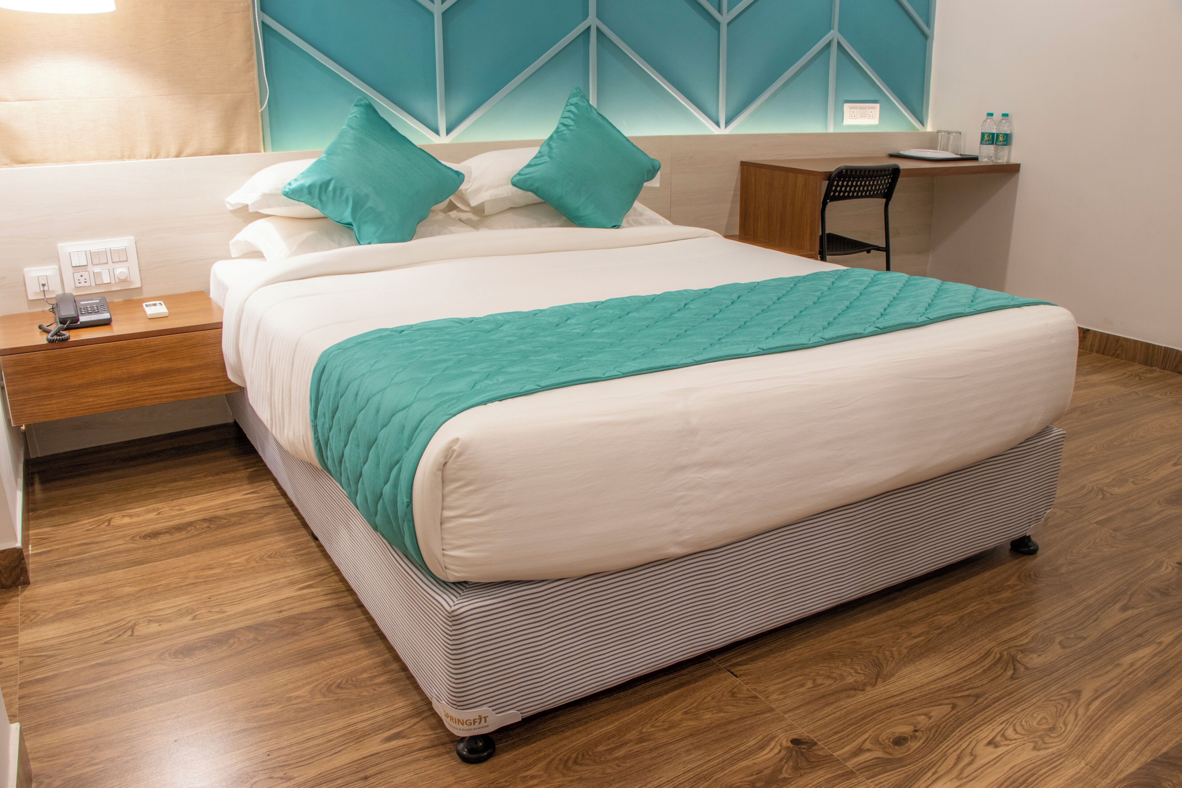 A hotel room with a bed, a desk, a chair, and a nightstand with a phone and a power outlet.