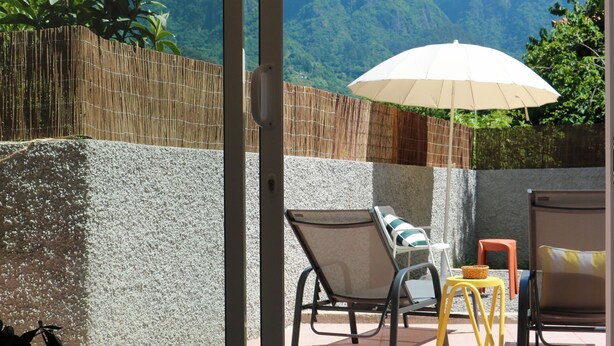 A room with a sliding glass door leading to an outdoor patio with a view of mountains, a chair, a small table, and an umbrella. - Araça Studio - Atlantic Mountain Living Surrounded by mountains and nature (São Vicente)