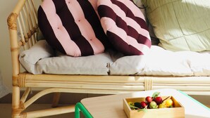 A wicker sofa with striped cushions, a green pillow, and a wooden side table with a fruit bowl. - Araça Studio - Atlantic Mountain Living Surrounded by mountains and nature (São Vicente)