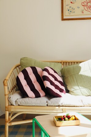 A wicker sofa with striped cushions, a green pillow, and a wooden side table with a fruit bowl. - Araça Studio - Atlantic Mountain Living Surrounded by mountains and nature (São Vicente)