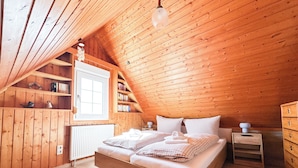 A wooden-paneled room with a bed, a nightstand, a dresser, and a shelf with books and decorative items.