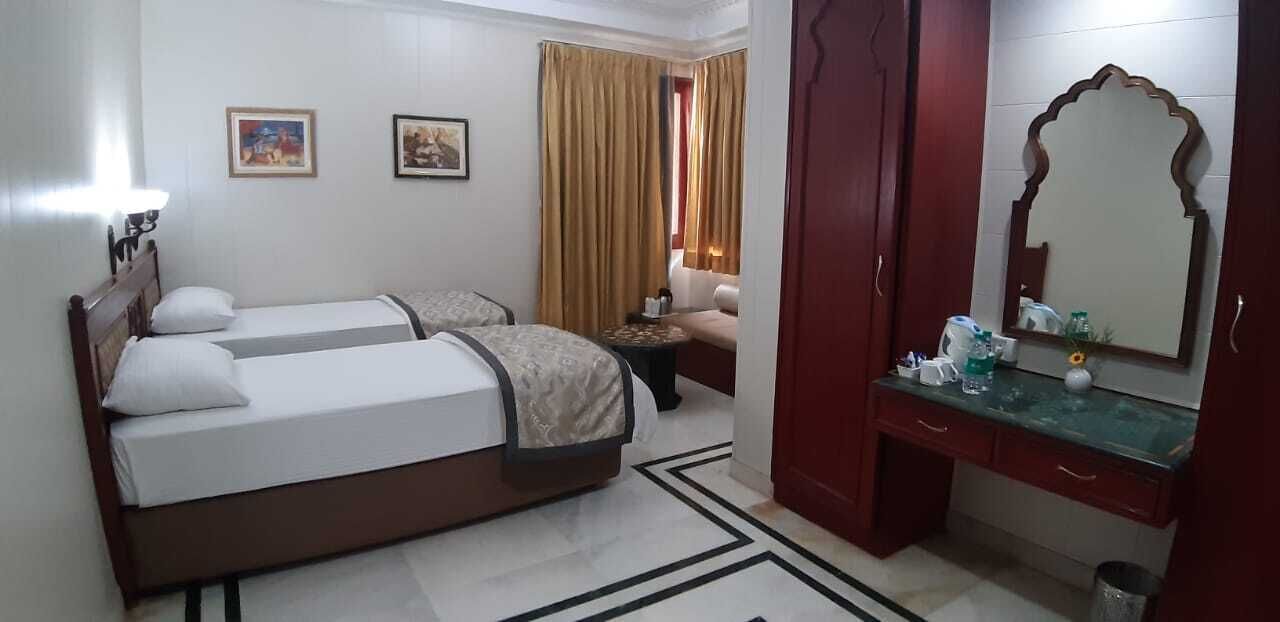Habitación de hotel con dos camas, zona de estar con un sofá, una mesita y un espejo, además de una ventana con cortinas.