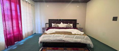 A bedroom with a bed, a nightstand, a bedside table, a patterned rug, and a window with curtains.