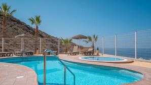 Piscina con sillones de descanso y sombrillas, rodeada de una valla y palmeras.