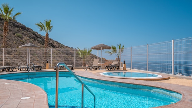 Une piscine entourée d’une clôture et de palmiers, avec des chaises longues et des parasols.