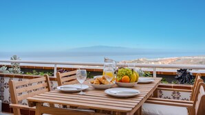Una mesa de madera con platos, una jarrón y un cuenco de fruta en un balcón con vista a una ciudad y una montaña.