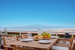 Ein Holztisch mit Tellern, einer Kanne und einer Obstschale auf einem Balkon mit Blick auf eine Stadt und ein Gebirge.