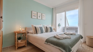 Un dormitorio con cama, una mesita de noche con lámpara, una ventana con cortinas y un muro con cuadros.