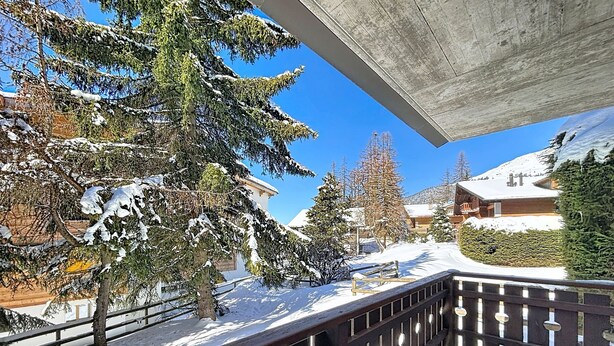 A snow-covered balcony with a wooden railing, overlooking a snowy landscape with trees and buildings. - A living room with dining area and fireplace, a separate kitchen with dishwasher, a double bedroom, (Verbier)