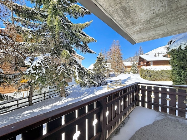 A snow-covered balcony with a wooden railing, overlooking a snowy landscape with trees and buildings. - A living room with dining area and fireplace, a separate kitchen with dishwasher, a double bedroom, (Verbier)
