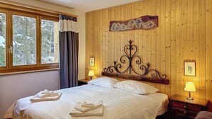 A bedroom with a bed, two bedside tables, a window with curtains, and a wooden wall with a decorative panel. - A living room with dining area and fireplace, a separate kitchen with dishwasher, a double bedroom, (Verbier)