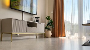 Ein modernes Wohnzimmer mit einem an der Wand befestigten Flachbildfernseher, einem schlichten Konsolentisch, einer Topfpflanze und großen Fenstern mit durchscheinenden Vorhängen.
