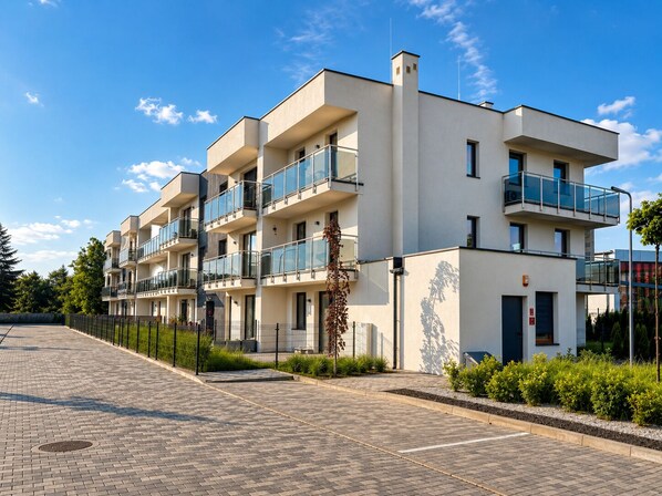 A modern multi-story residential building with balconies, a paved driveway, and a clear sky.