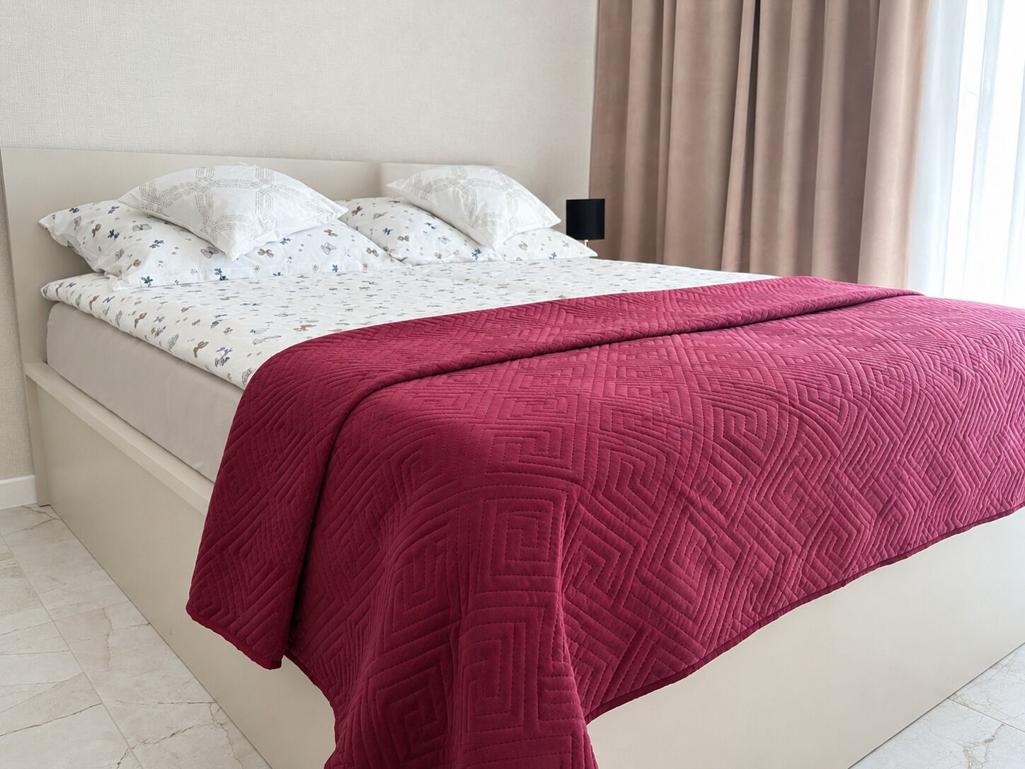 A bed with a patterned bedspread and pillows, a bedside lamp, and a window with curtains.