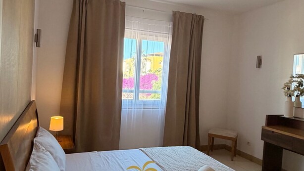 A hotel room with a bed, a bedside table with a lamp, a window with curtains, a small bench, and a vanity with a mirror. - Comfy 2-bedroom apartment with AC in Santa Maria (Santa Maria)