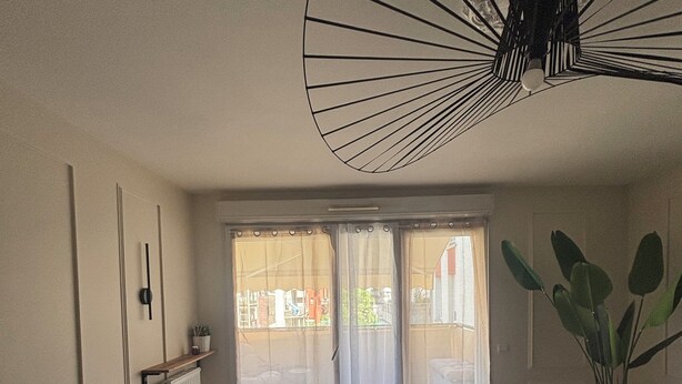 Una habitación con un ventanal grande, un ventilador de techo, un radiador, un sofá verde, una planta en maceta y una alfombra.