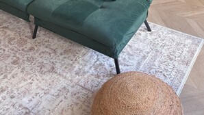 Un sofá verde con patas negras, un tapete estampado y un taburete tejido.