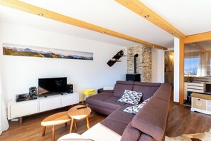 A modern living room with a large brown sofa, wooden flooring, a mounted TV, and a stone accent wall. - Panoramic Mountain Views, Spacious Sauna, Dogs Welcome, Alpenjuwel 2 Oberstdorf (Oberstdorf)