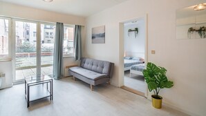 Una sala moderna con un sofá, una mesita de vidrio, un ventanal grande, una cama y una planta en maceta.