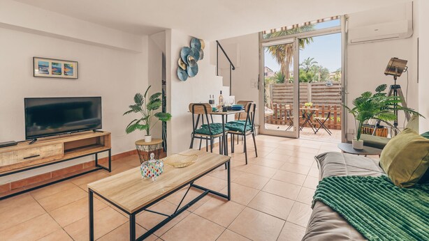 Ein Wohnzimmer mit Fernseher, Couchtisch aus Holz, grünem Sofa und einem Essbereich mit Stühlen und Tisch.