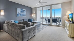 Ein Wohnzimmer mit grauem Sofa, Deckenventilator und großem Fenster mit Blick auf die Stadt.