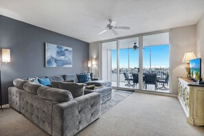 A living room with a grey sofa, a ceiling fan, and a large window with a view of the city. - Regatta - Walk to the Beach! (Naples)
