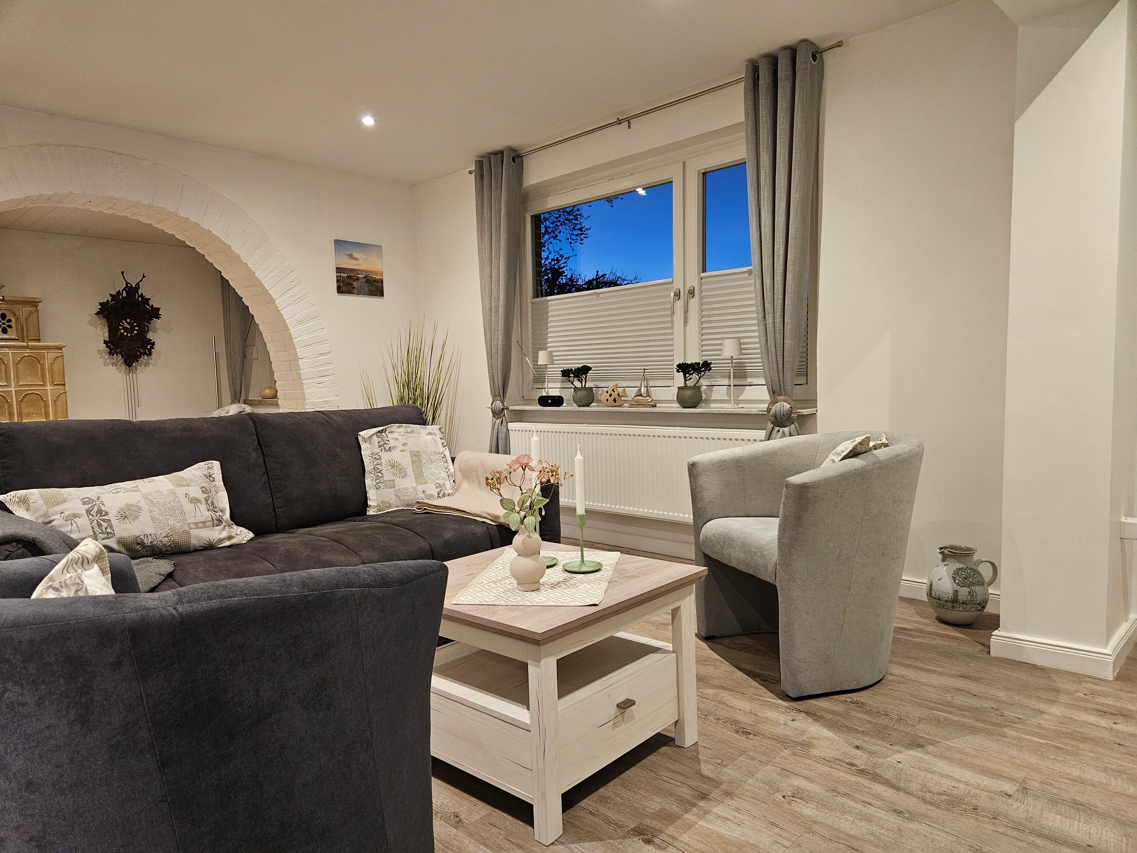 Uma sala de estar moderna com um sofá cinzento, uma mesa de centro e uma poltrona cinzenta. Há uma janela com cortinas, um radiador e vista para o exterior.