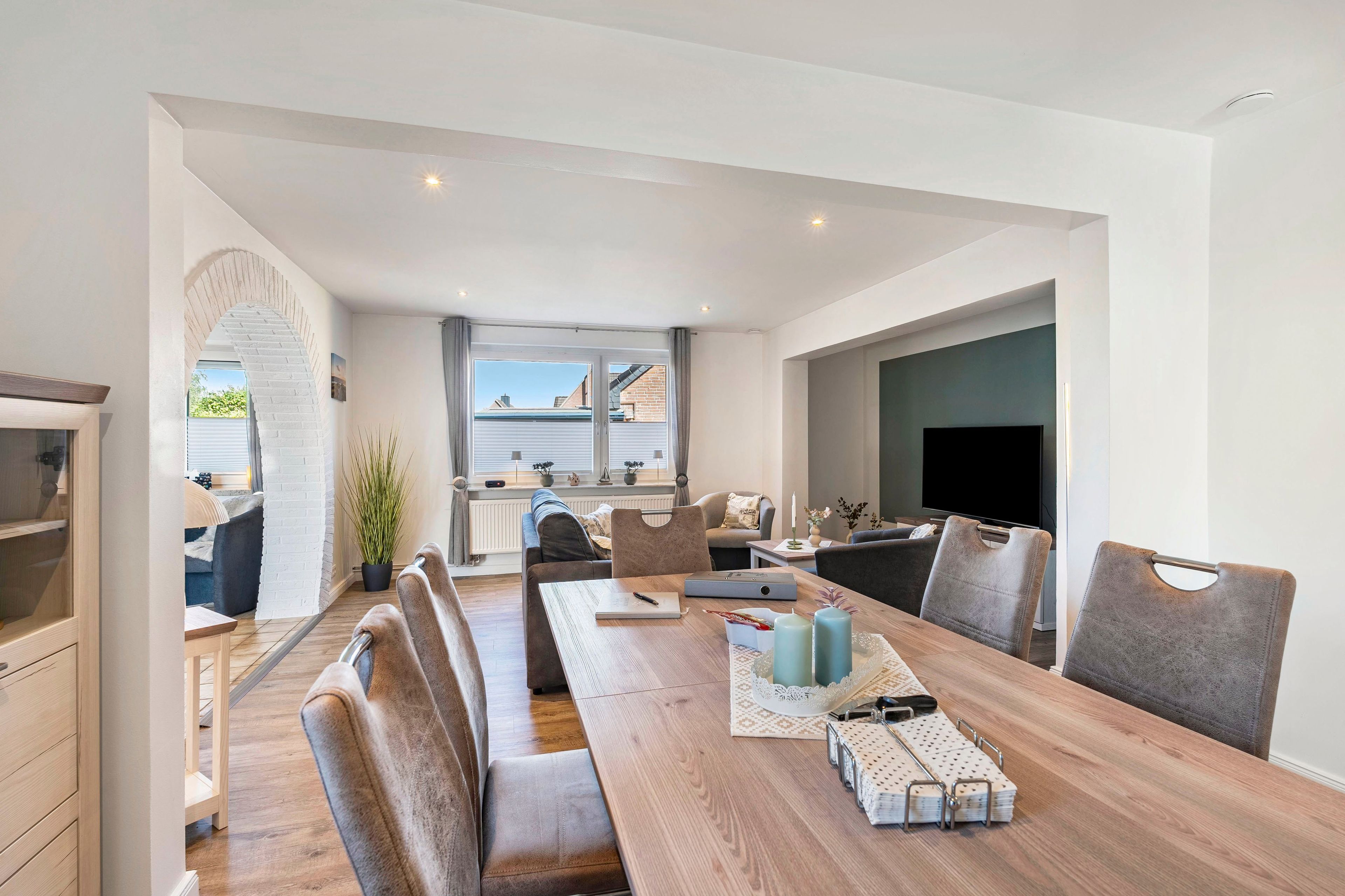 A modern living room with a wooden dining table, cushioned seating, a flat-screen TV, and a view of the outside.