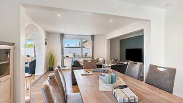 A modern living room with a wooden dining table, cushioned seating, a flat-screen TV, and a view of the outside.
