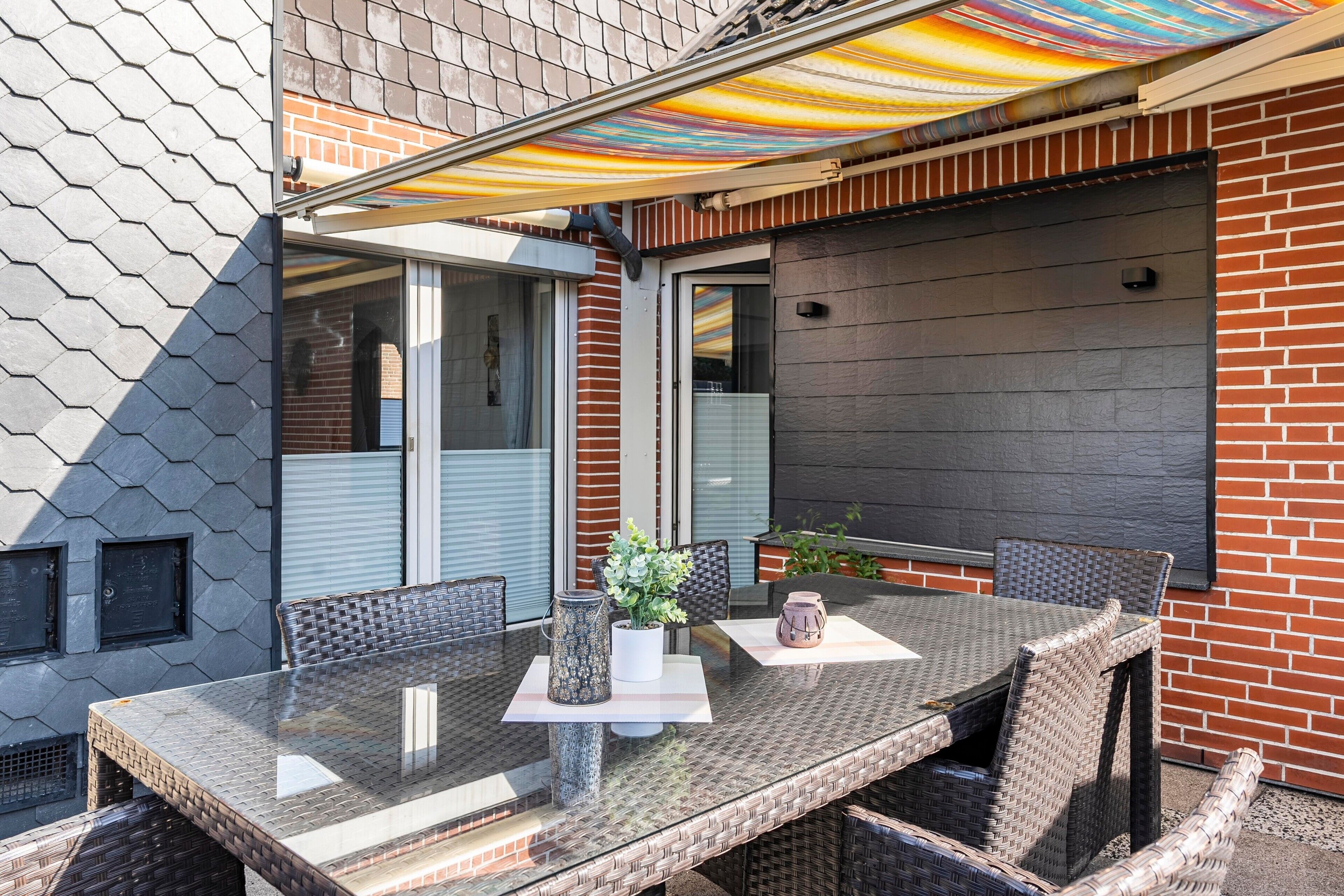 En udendørs terrasse med et glasbord, fletstole og en farverig markise.