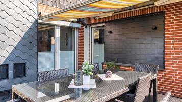 En udendørs terrasse med et glasbord, fletstole og en farverig markise.