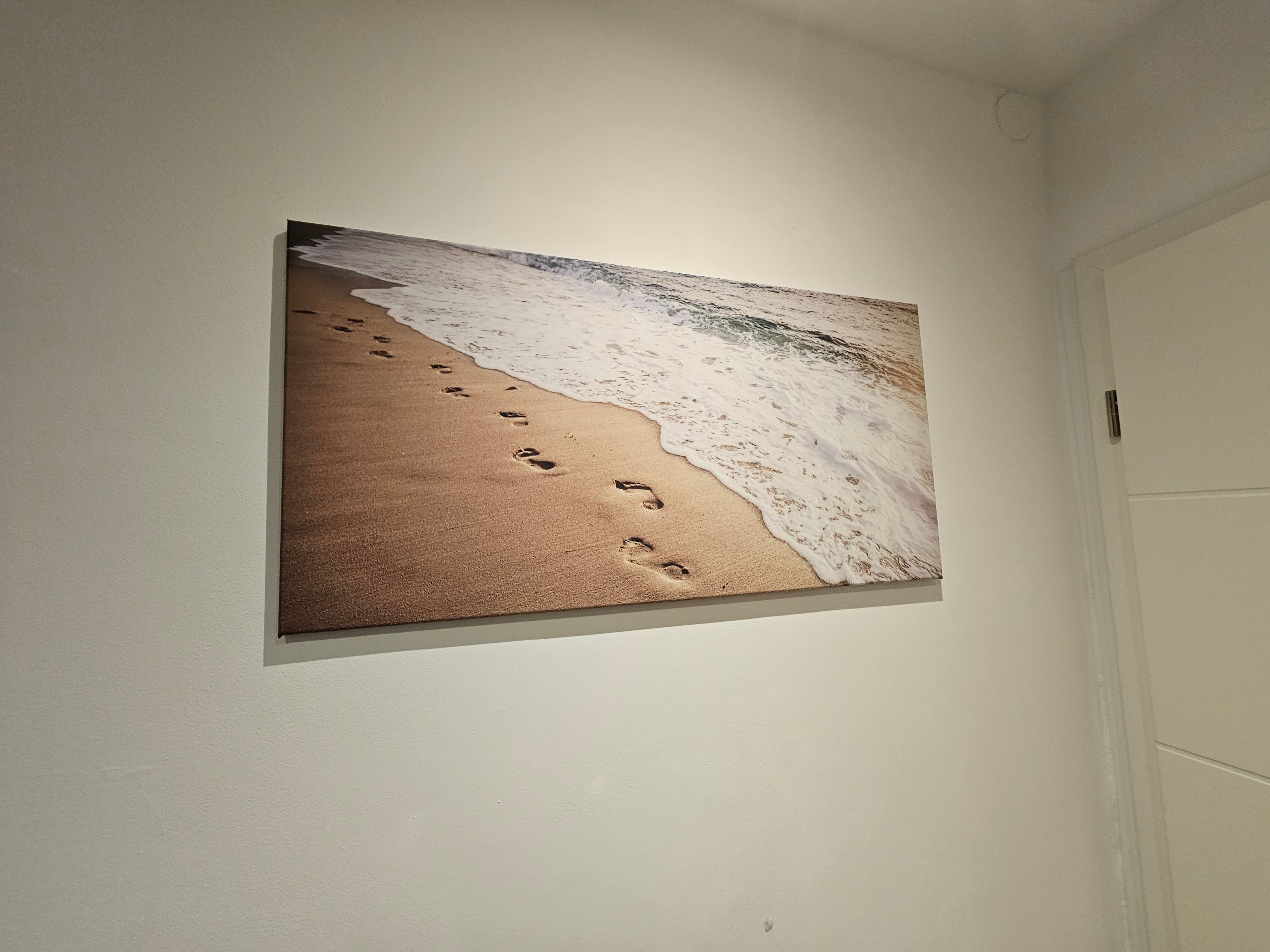 En inramad tavla som föreställer en strandscen med fotspår som leder ner mot vattnet.