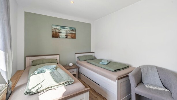A bedroom with two beds, a nightstand, a chair, and a painting on the wall. - Holiday Apartment Elisabeth at Lake Plön (Ascheberg (Holstein))