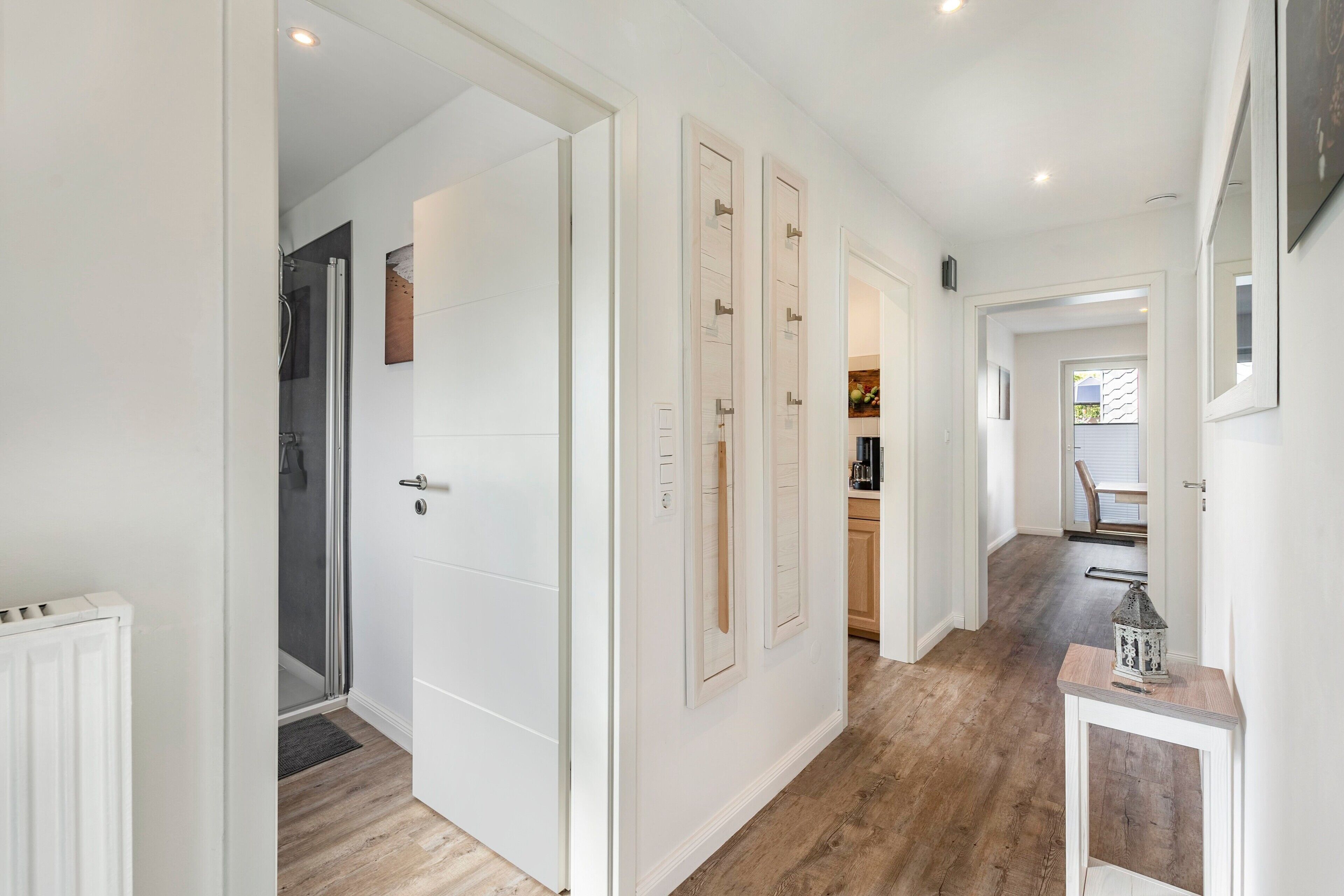 A hallway with white doors, a shower area, and a kitchen visible through an open door.
