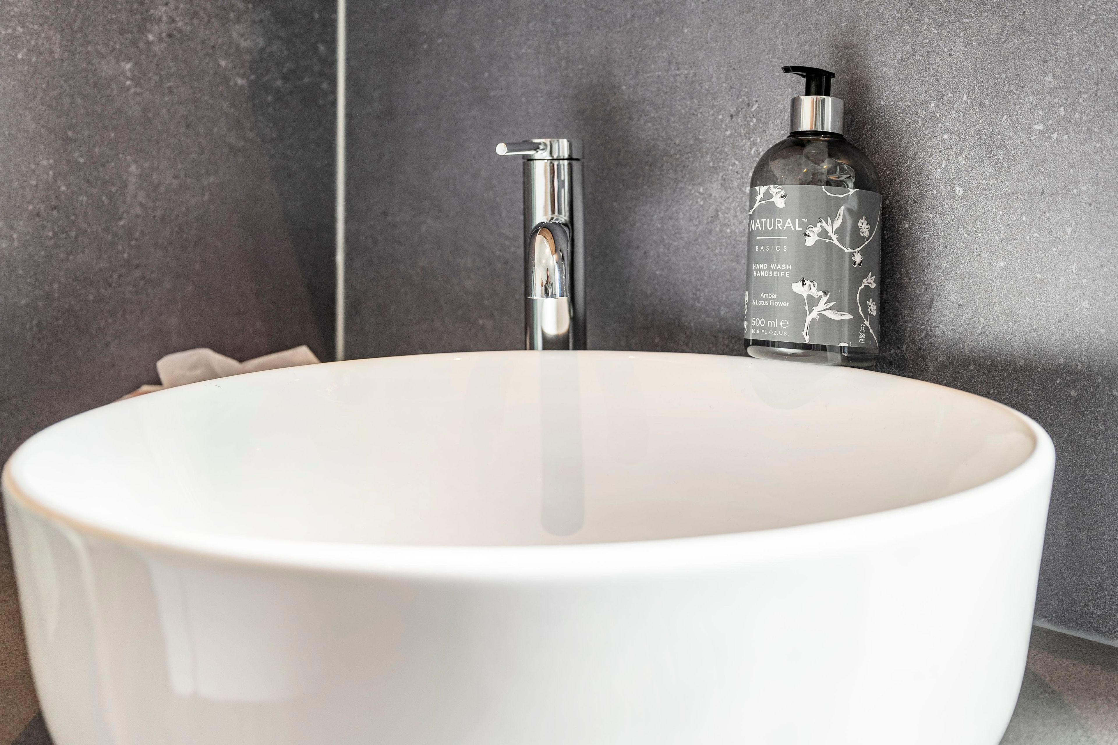 Ein weißes Waschbecken mit einem modernen Wasserhahn und einer Seifenspenderflasche an einer strukturierten Wand.