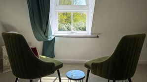 Ein Zimmer mit einem großen Bogenfenster, zwei grünen Stühlen, einem kleinen runden Tisch und einer weißen Wand.