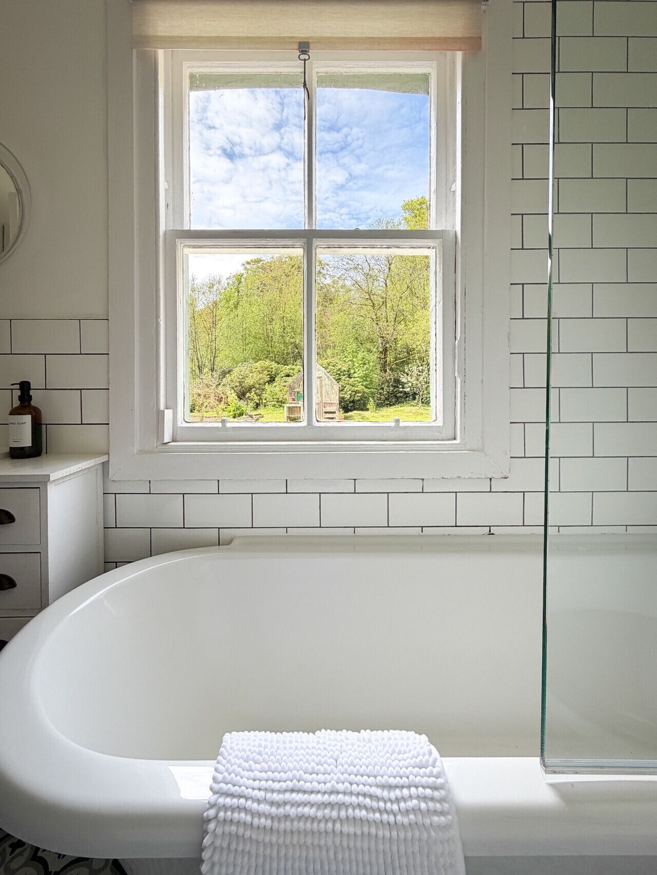 A bathroom with a white bathtub, a window with a view of trees, and a white tiled wall.