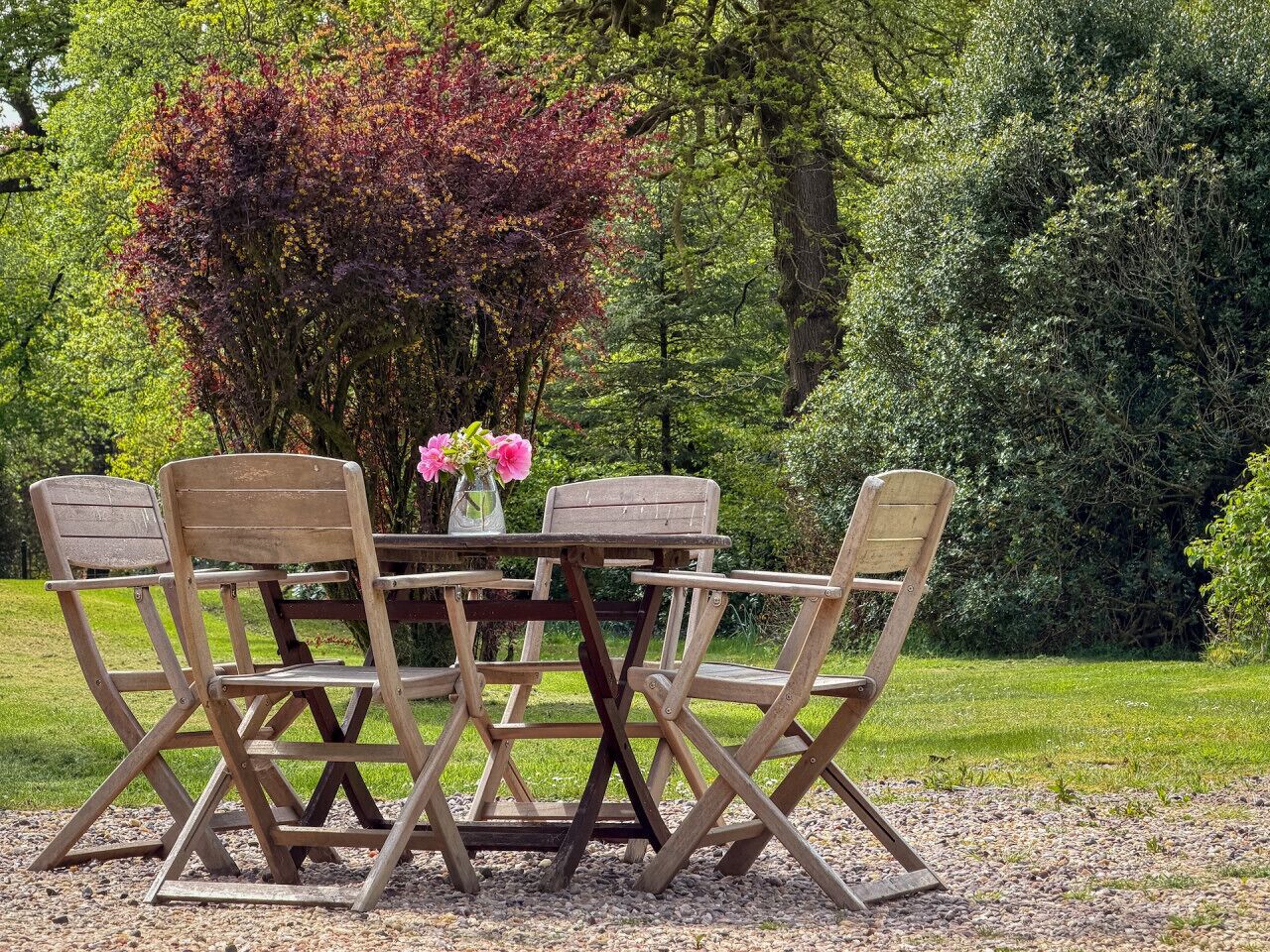 Een houten tuinmeubelset met tafel en stoelen, geplaatst op een grindpad, met een bloevas.