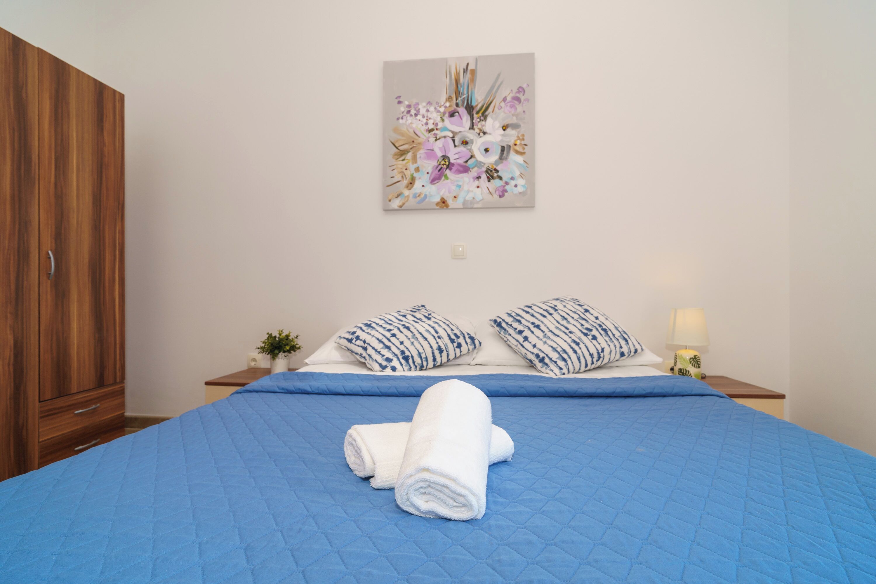 A bedroom with a blue quilted bedspread, two patterned pillows, a rolled white towel, a wooden wardrobe, a small plant, and a wall-mounted painting.