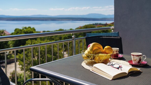 A balcony with a table set for breakfast, a book, and a view of a lake and mountains. - Apartment for 4 people in Dramalj with sea view & parking space (Dramalj)