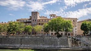 Un edificio histórico de varios pisos, una torre destacada y un río al frente.
