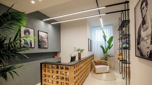 A reception area with a wooden reception desk, a white sofa, potted plants, framed pictures on the wall, and a staircase with a black railing. - Daplace - LE SUITES DEGLI ALTOVITI (Rome)