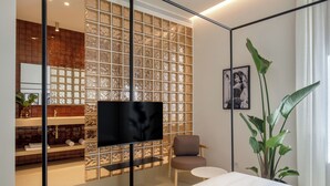 A modern hotel room with a large TV, a small round table, a comfortable chair, a large plant, and a bathroom with a tiled backsplash. - Daplace - LE SUITES DEGLI ALTOVITI (Rome)