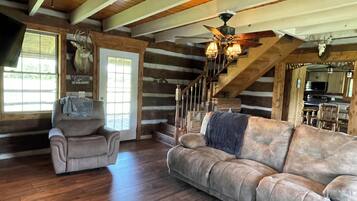 A log cabin interior with a wooden staircase, a sofa, a chair, and a ceiling fan.