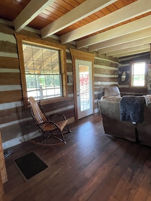 L’intérieur d’une cabane en rondins, avec un fauteuil à bascule, un canapé et une porte donnant sur l’extérieur.