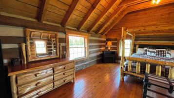 A log cabin interior with a wooden dresser, a bed with a headboard, a fireplace, and a window.