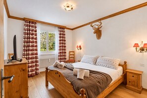 Un dormitorio con una cama de madera, un escritorio con computadora, una ventana con cortinas, un adorno de cabeza de ciervo montado en la pared y una mesita de noche con lámparas.